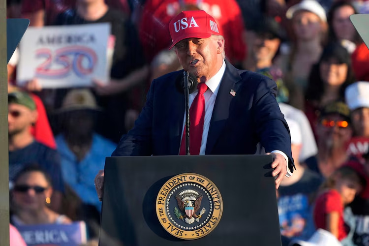 Donald Trump à la tribune en tant que président des États-Unis, il porte une casquette rouge USA, une foule est visible en arrière plan.