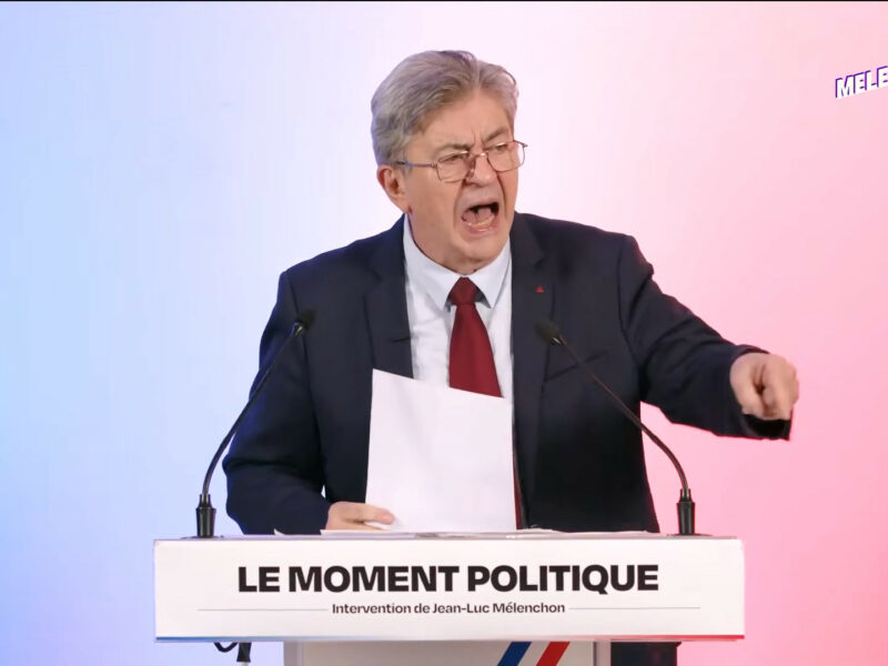 Jean-Luc Mélenchon, en costume sombre et cravate rouge, crie, une feuille à la main et l’index pointé vers le public, derrière un pupitre marqué « Le moment politique – Intervention de Jean-Luc Mélenchon ». Fond dégradé bleu, blanc, rouge, logo « Melenchon2027.fr » en haut à droite.