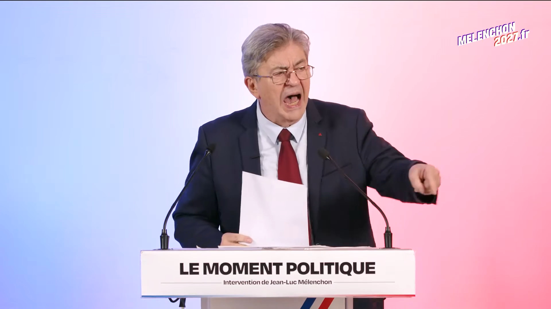 Jean-Luc Mélenchon, en costume sombre et cravate rouge, crie, une feuille à la main et l’index pointé vers le public, derrière un pupitre marqué « Le moment politique – Intervention de Jean-Luc Mélenchon ». Fond dégradé bleu, blanc, rouge, logo « Melenchon2027.fr » en haut à droite.